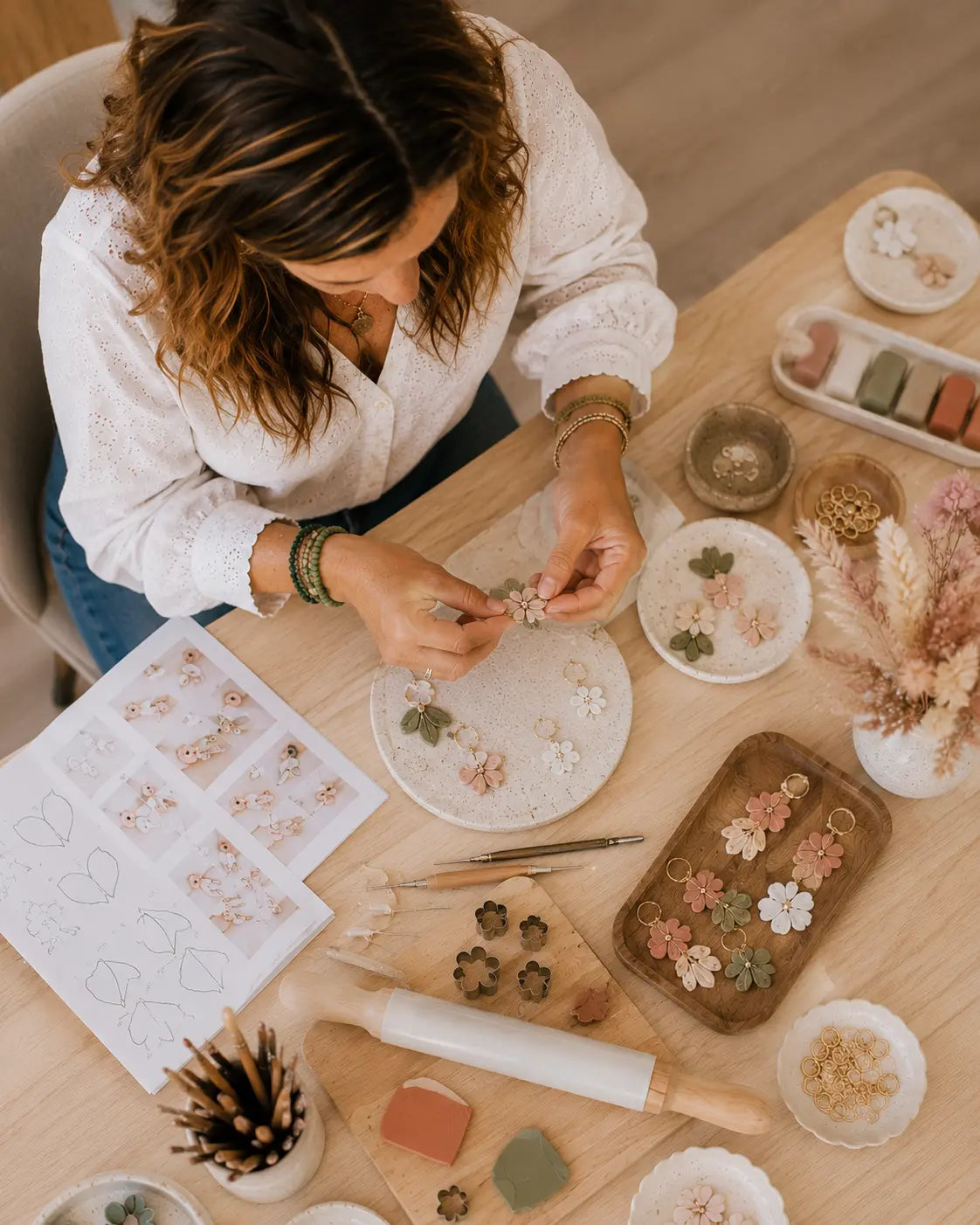 A woman in a white blouse sits at a wooden table, crafting delicate floral decorations with small pieces of fabric, beads, and tools spread out on the table surface.